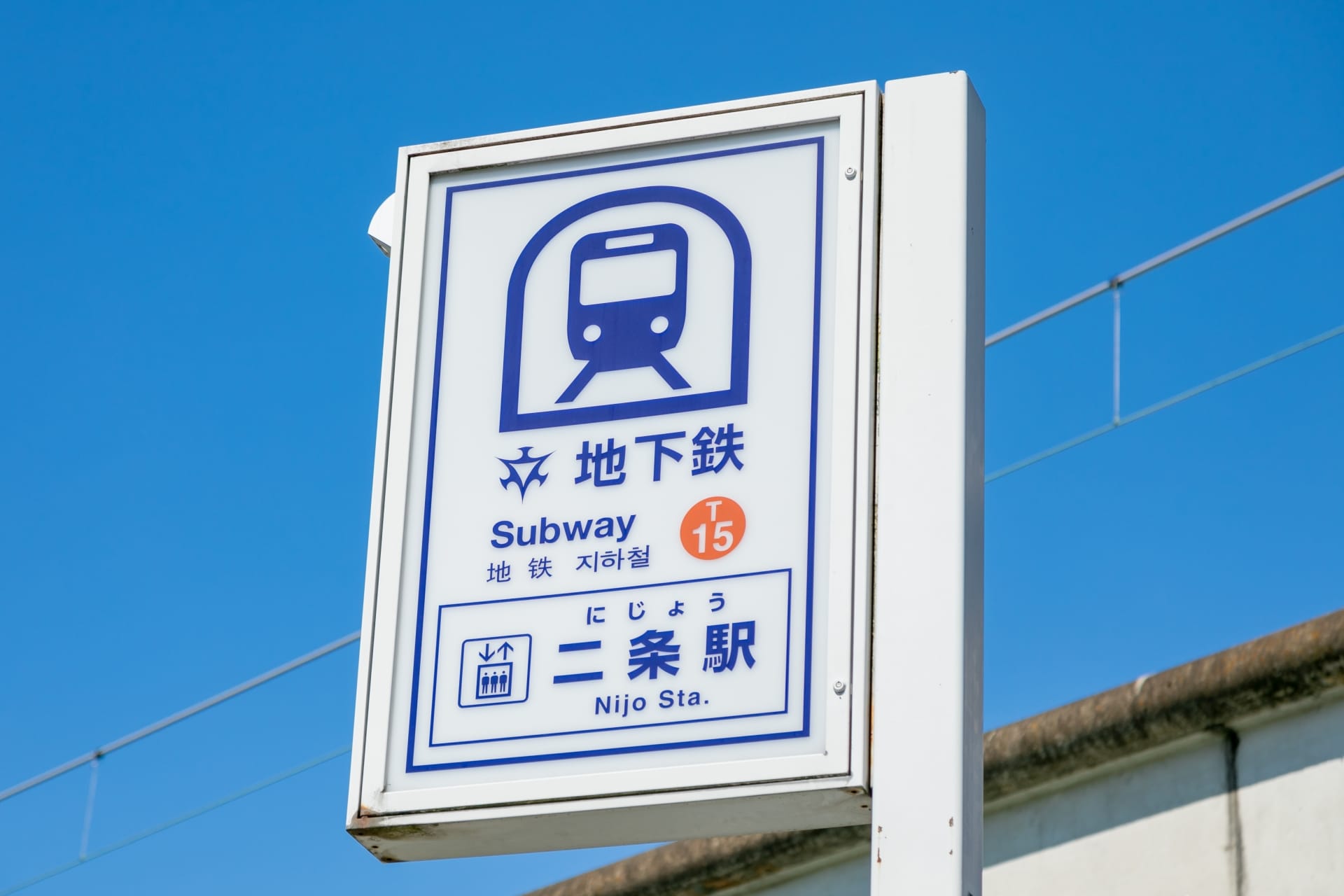 地下鉄のアイコンと日本語、英語、フランス語教室マンツーマンの文字が描かれた二条駅の標識が、澄んだ青空に堂々と立っています。.