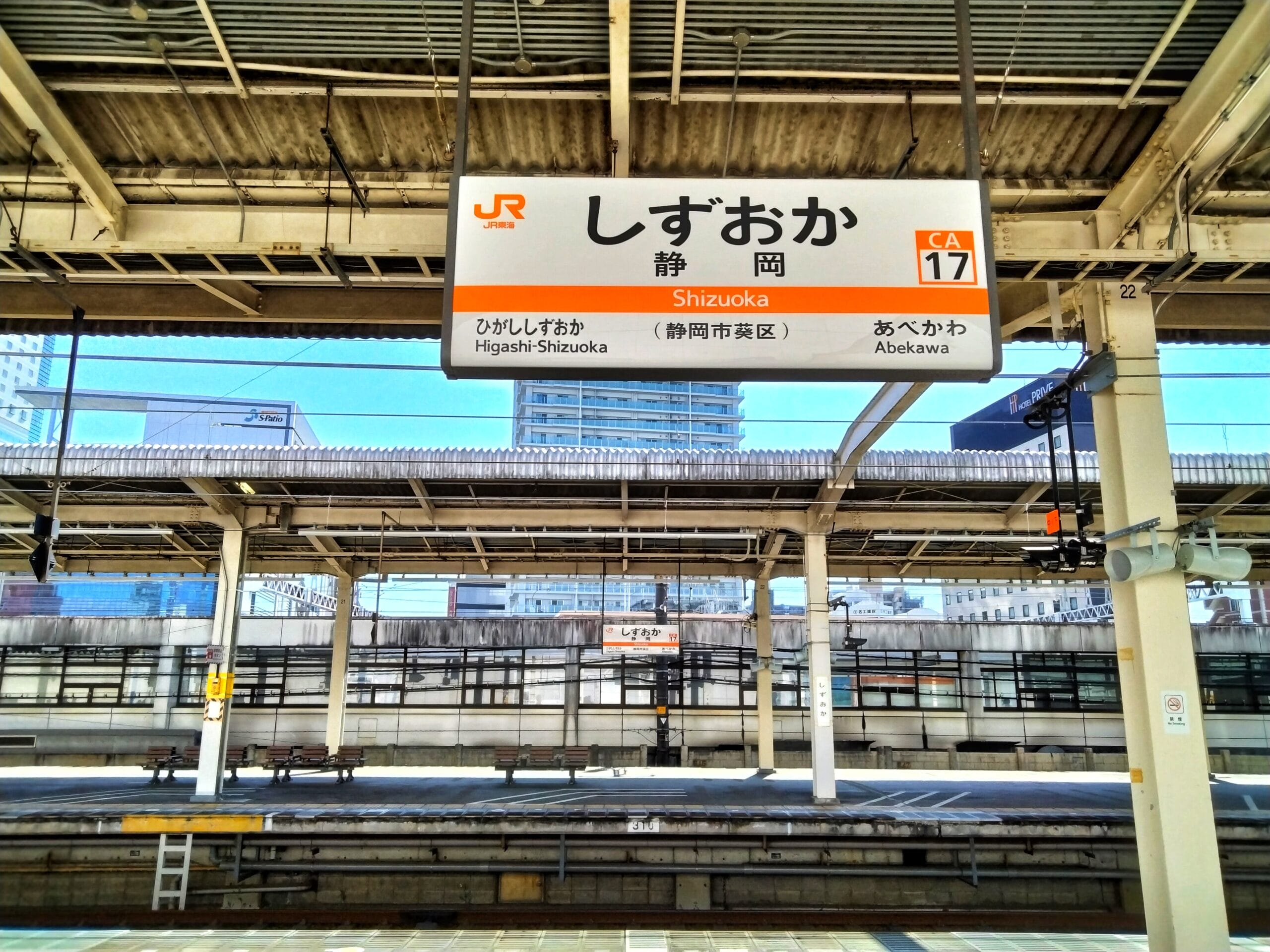 日本の静岡駅の空の電車のプラットホーム。頭上には日本語と英語の標識があります。静岡・三島が地域の魅力を添え、背景には優雅な建物がそびえ立っています。.