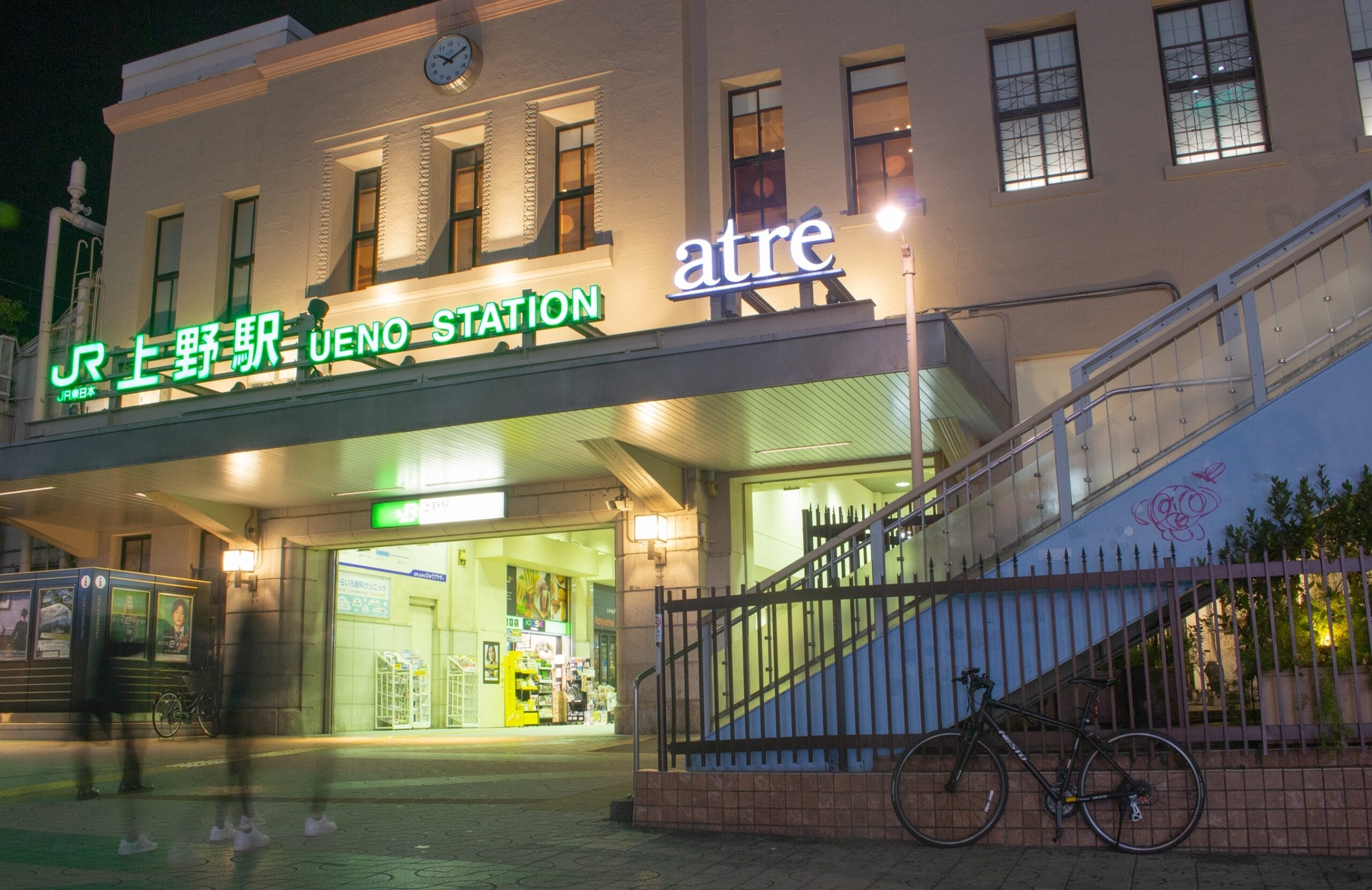 上野駅入口の夜景。明るい看板と柵のそばに駐輪された自転車。ぼやけた人々が通り過ぎ、動きを感じさせます。.