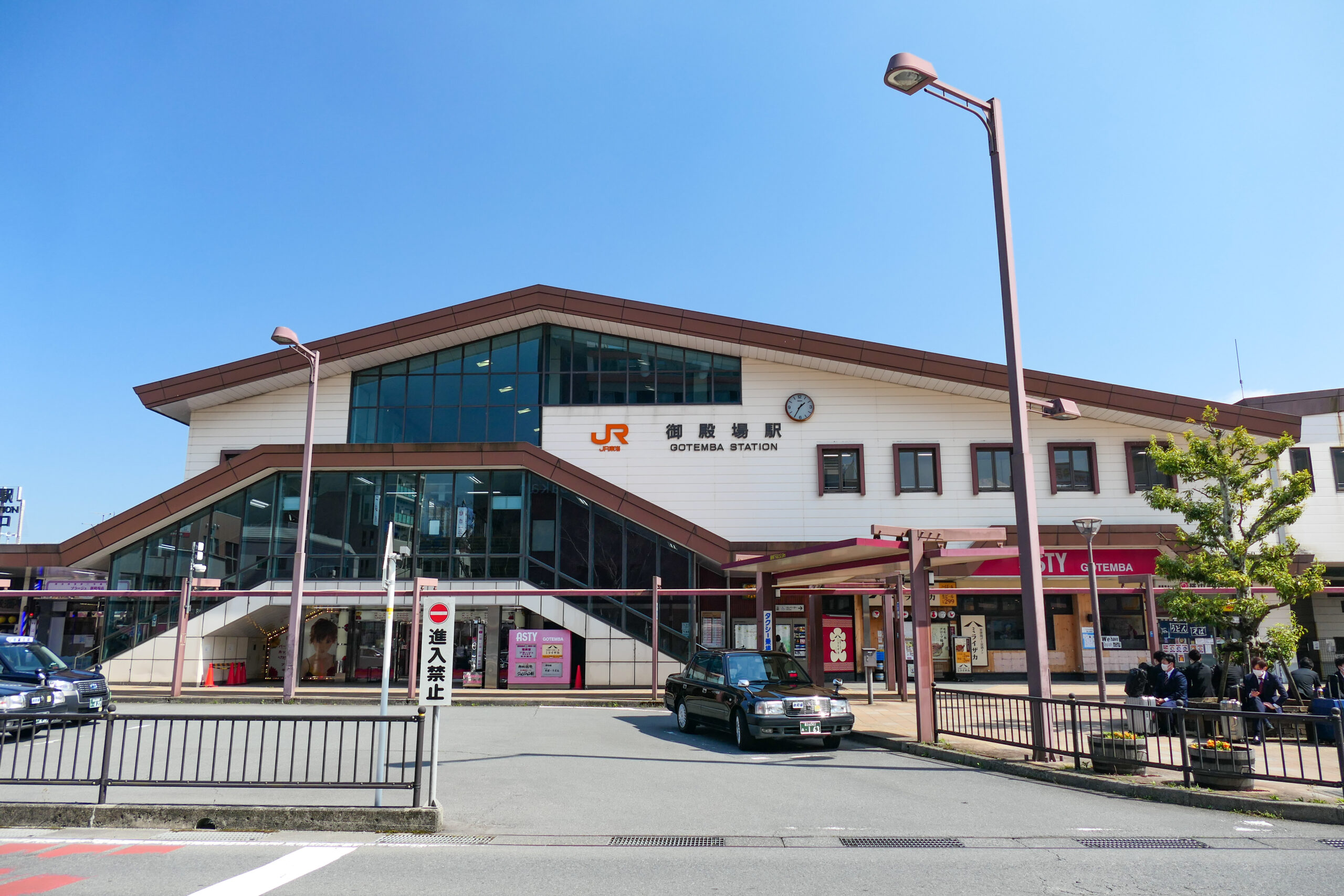 日本のJR大津駅の外観。晴れた日の駅舎、入口、停車中のタクシー、そして正面入口付近に集まる人々の様子が写っている。遠くには「フランス語教室」の看板が見え、活気あふれる雰囲気に国際的な趣を添えている。.