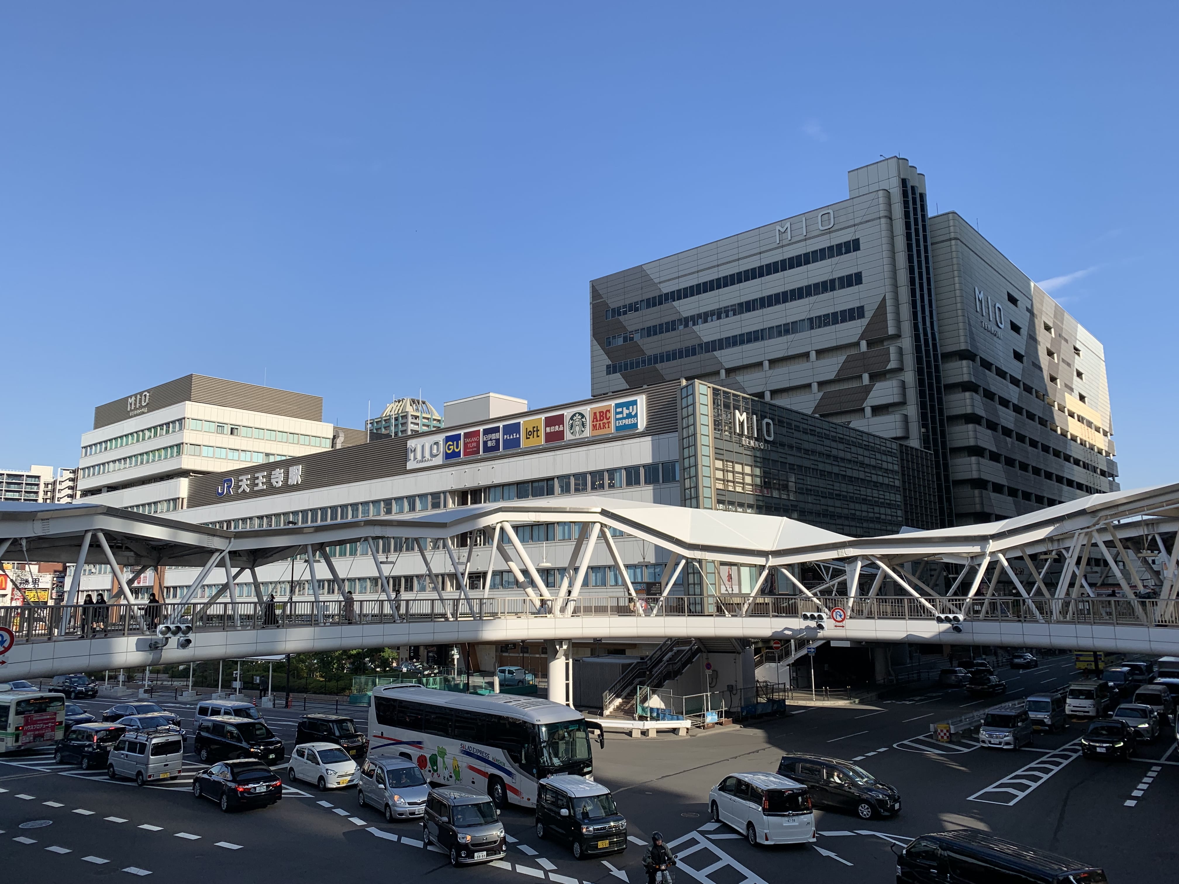 晴れ渡った青空の下、「MIO」の看板、高架の歩行者通路、交通量の多い近代的な複合商業施設。.