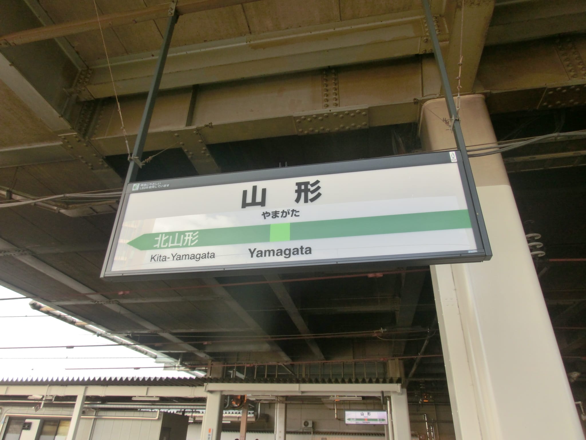 「山形」と書かれた駅名標と北山形行きの案内。背景には建物と線路が見え、山形・米沢らしさを感じさせる。.
