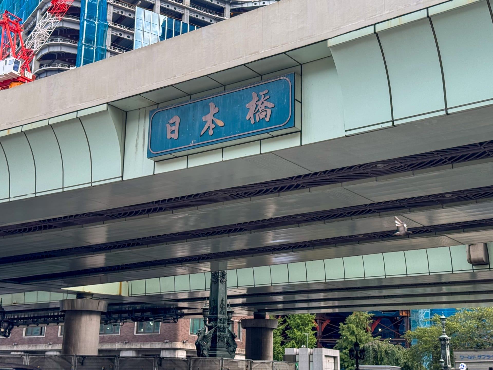 日本橋の近くには日本語の文字で書かれた標識のある高架道路が伸びており、背景には建設中の建物、その下には生い茂る木々が見える。.