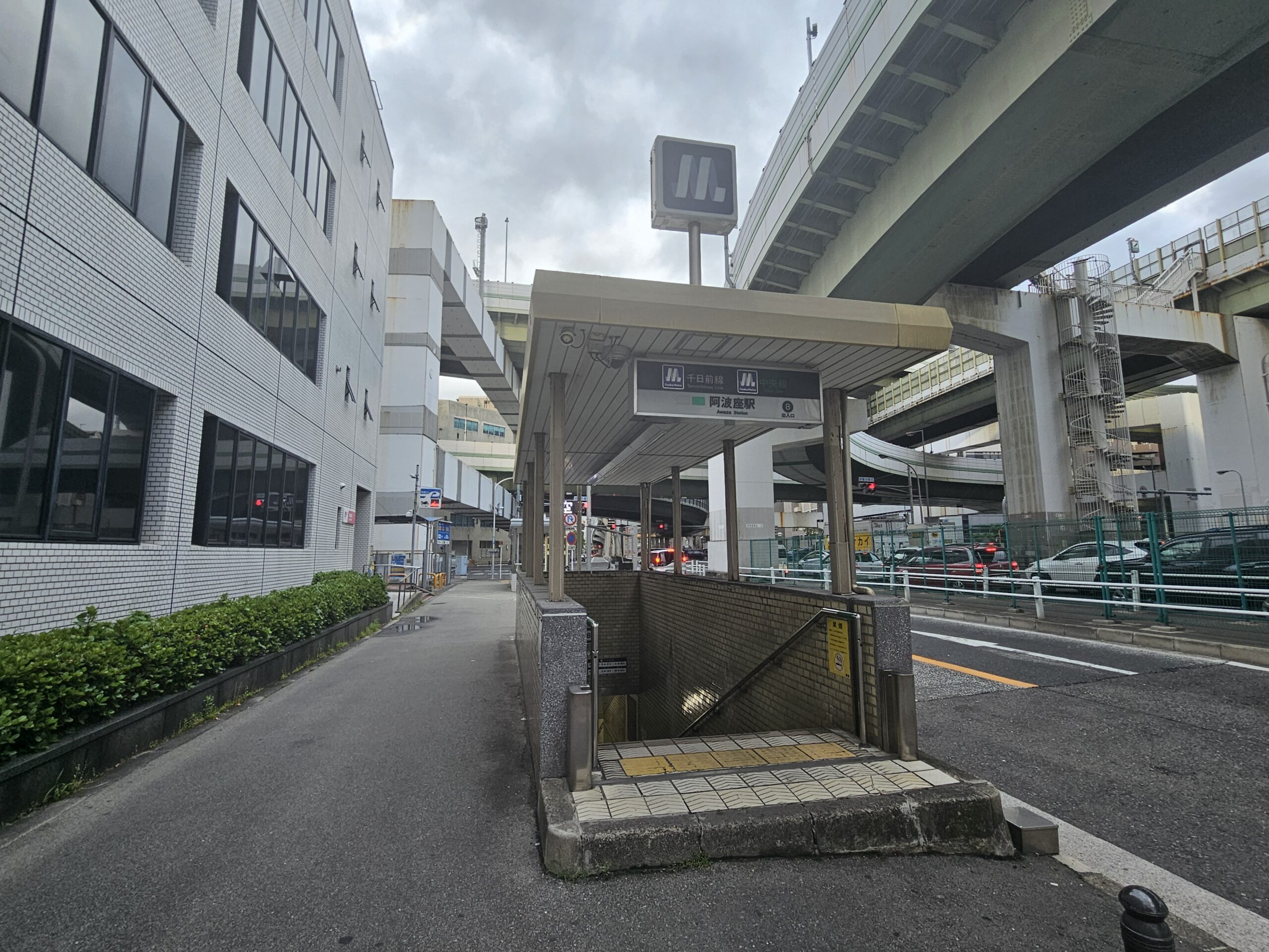 阿波座近くの市街地にある地下鉄駅の屋根付き入口。周囲は建物に囲まれ、空は曇り、頭上には高架道路が走っている。.