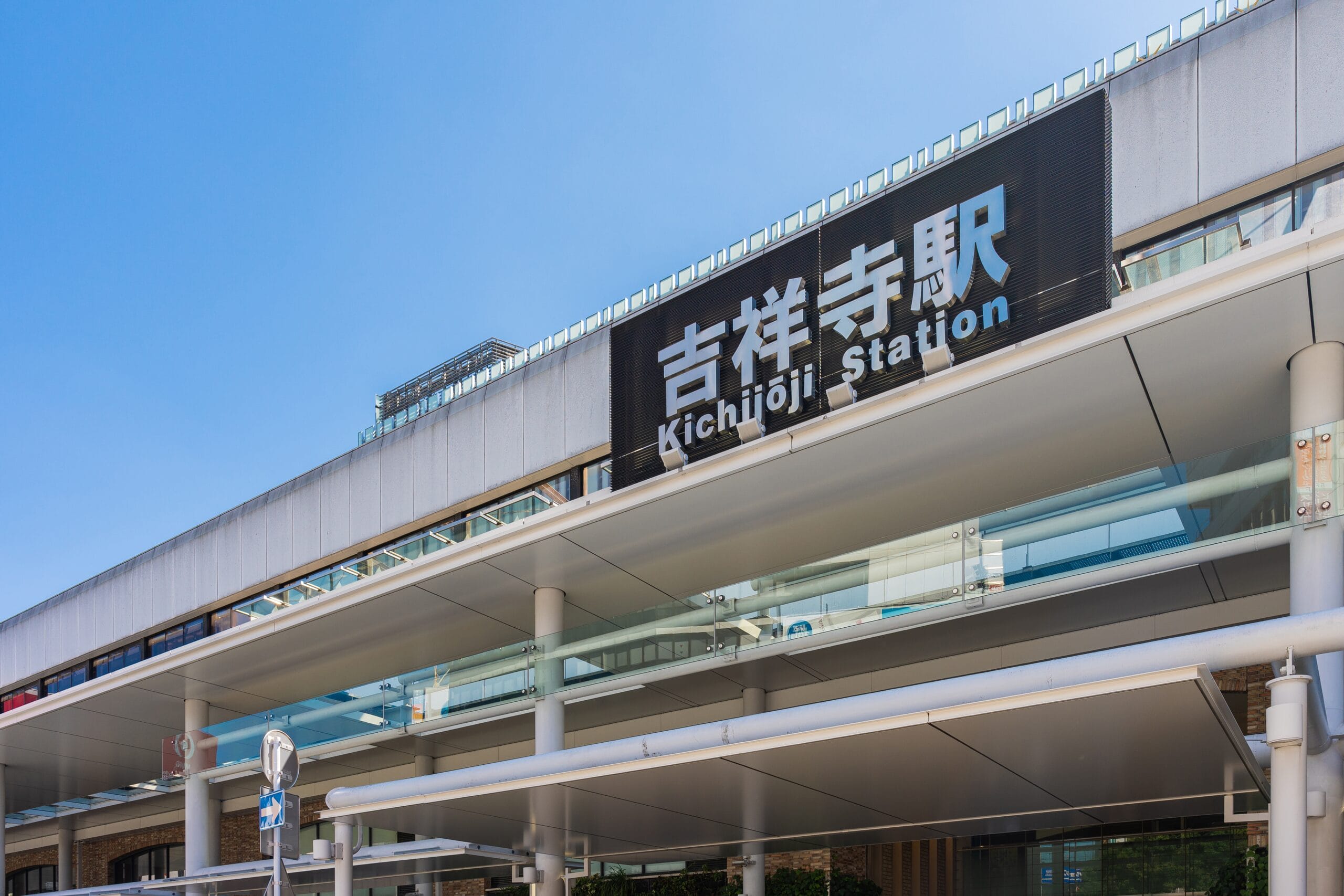 日本の吉祥寺駅の外観。青空の下、入口の上にある看板に日本語と英語で駅名が記されている。.