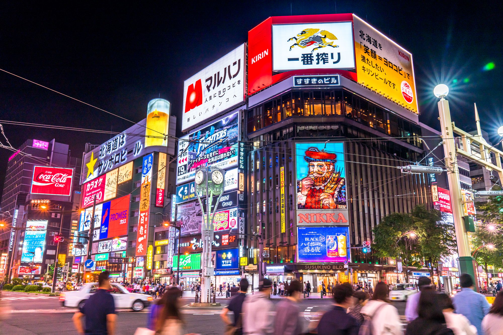 夜、色とりどりのイルミネーションが輝く看板やネオンサインが立ち並び、通りを人々が歩く、にぎやかな都市の交差点。.